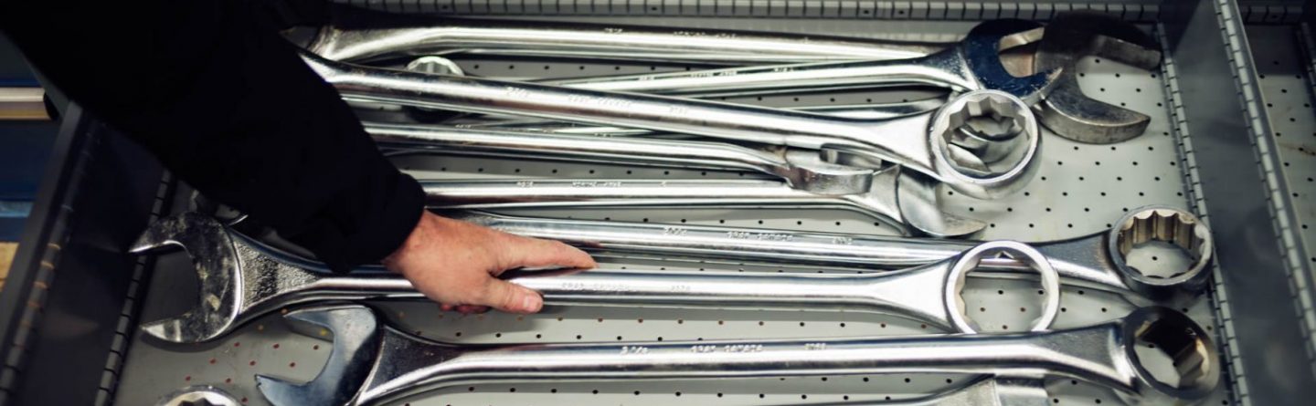 A person’s hand reaching for a large metal wrench among several oversized wrenches arranged in a drawer with a perforated metal bottom.
