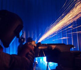 A worker wearing a protective welding helmet and reflective vest uses a grinder on metal, creating bright orange sparks in a dimly lit industrial setting with blue lighting.