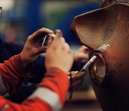 Two people in safety gear inspect a large industrial metal part; one holds a tool to the surface while the other takes a close-up photo, capturing details for documentation.