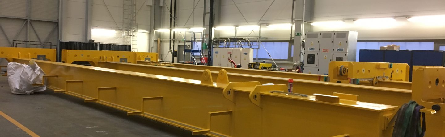 A large yellow metal crane girder is resting on the floor of an industrial warehouse, surrounded by tools, equipment, and pallets. The space is well-lit with high ceilings and control panels in the background.