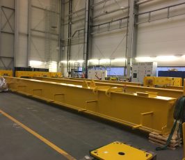 A large yellow metal crane girder is resting on the floor of an industrial warehouse, surrounded by tools, equipment, and pallets. The space is well-lit with high ceilings and control panels in the background.