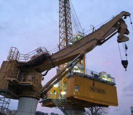 A large yellow Palfinger crane with an extended articulated arm stands at a construction site during dusk, illuminated by work lights, with buildings visible in the background.
