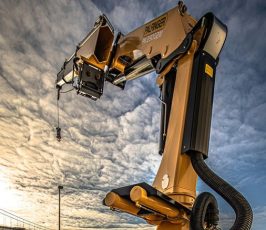 A large yellow industrial robotic arm extends upwards outdoors under a dramatic cloudy sky, with a bridge and water visible in the background.