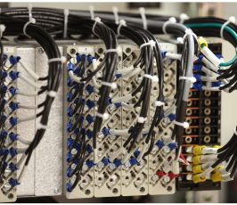 Close-up of a network server panel with multiple black cables connected to labeled ports, secured with white cable ties, showing organized wiring in a data center or telecom setup.