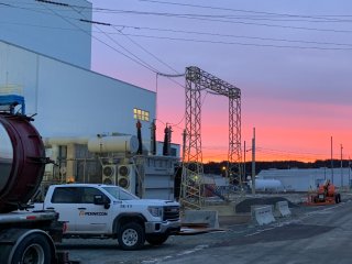 A construction site at sunset, featuring a white PennEcon truck, industrial equipment, and large building structures. The sky is vibrant with pink and purple hues.
