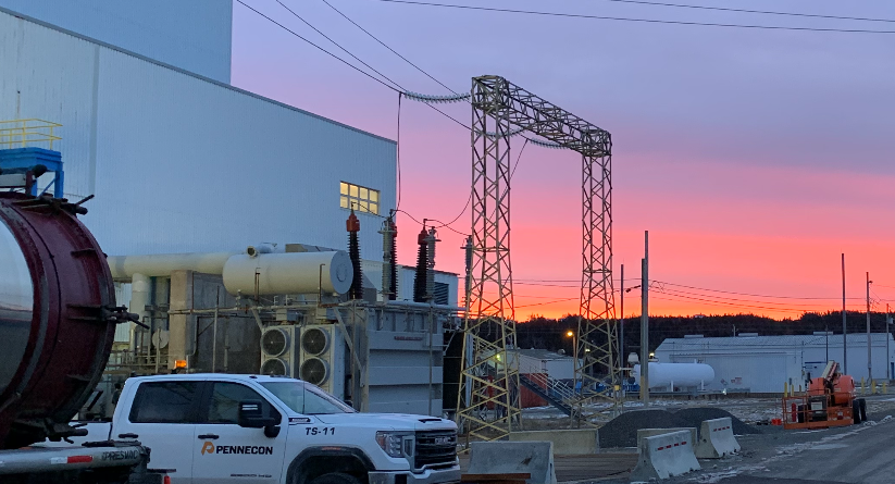 A construction site at sunset, featuring a white PennEcon truck, industrial equipment, and large building structures. The sky is vibrant with pink and purple hues.