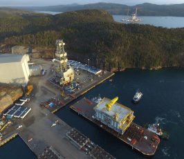 Aerial view of a waterfront industrial site with large structures, cranes, docked vessels, and a forested landscape surrounding a bay. An oil platform is visible in the water near the distant shoreline.