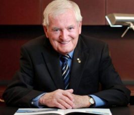 An older man with white hair, wearing a dark suit and striped tie, sits at a desk smiling with an open book in front of him. A lamp and brown shelves are visible in the background.