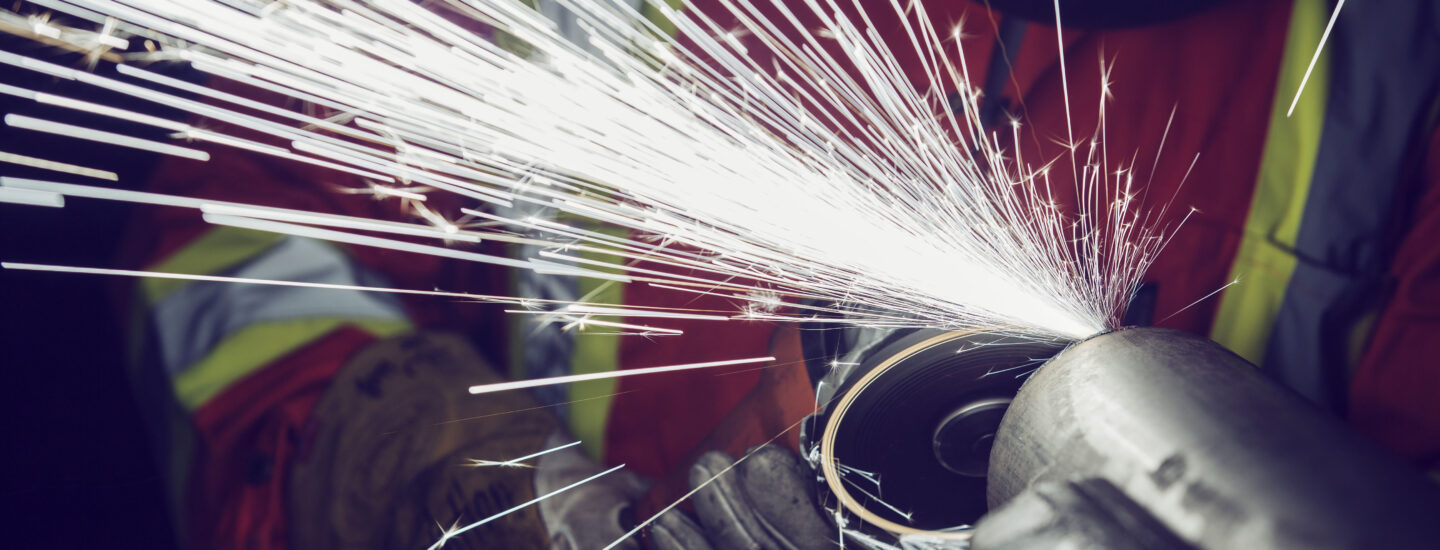 A worker wearing safety gear uses an angle grinder on metal, producing bright, flying sparks against a dark background.