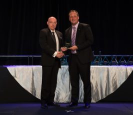 Two men in suits shake hands on a stage during an award ceremony, with trophies visible on a table behind them and a dark backdrop.