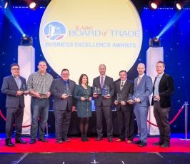 Eight people stand on a red carpet stage holding awards in front of a large sign that reads "St. John