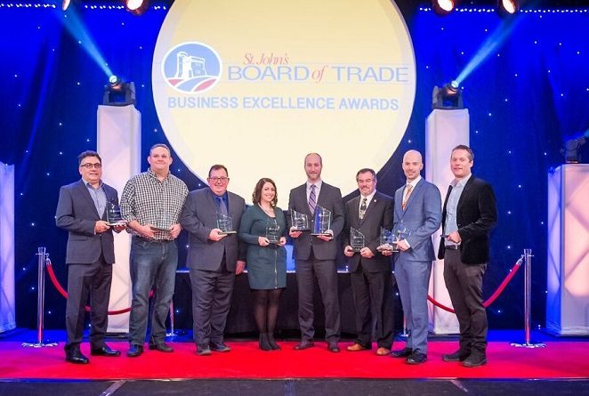 Eight people stand on a red carpet stage holding awards in front of a large sign that reads "St. John