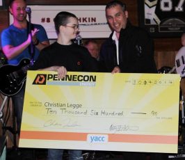 Two people stand holding an oversized check for $10,600 made out to Christian Legge from Pennecon Energy, dated 30/04/2014. A band and other people are visible in the background, and one man is clapping.