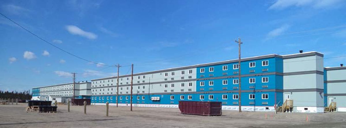 A long, rectangular, three-story blue and gray building sits on a dirt lot under a blue sky, with utility poles, a dumpster, and construction materials scattered nearby.