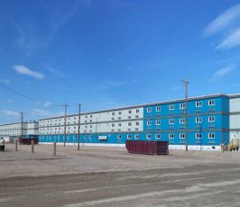 A long, rectangular, three-story blue and gray building sits on a dirt lot under a blue sky, with utility poles, a dumpster, and construction materials scattered nearby.