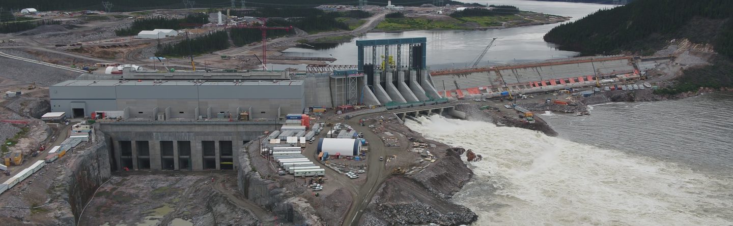 Aerial view of a large hydroelectric dam and power station under construction, with turbulent water flowing through spillways, surrounded by rocky terrain, equipment, and buildings, set in a forested landscape under a cloudy sky.