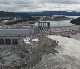 Aerial view of a large hydroelectric dam and power station under construction, with turbulent water flowing through spillways, surrounded by rocky terrain, equipment, and buildings, set in a forested landscape under a cloudy sky.