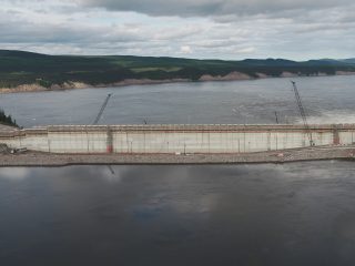 A wide concrete dam under construction spans a river, with cranes positioned along its length. The river is surrounded by forested hills and cloudy skies in the background.