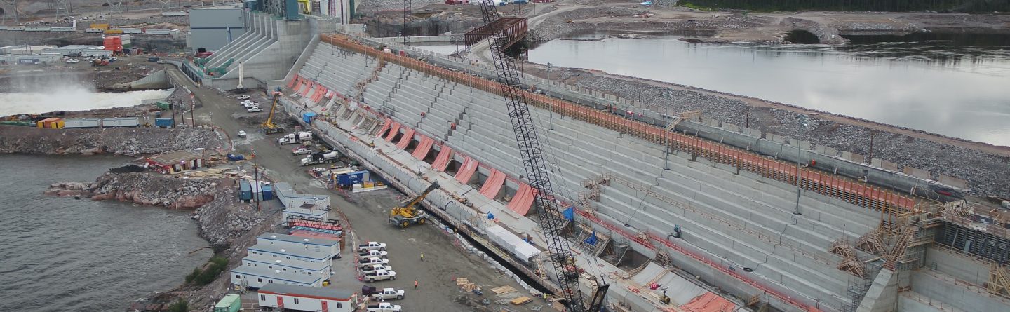 A large hydroelectric dam under construction, with cranes, vehicles, and equipment along the site; water on one side and construction trailers and buildings nearby under a cloudy sky.