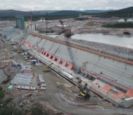 A large hydroelectric dam under construction, with cranes, vehicles, and equipment along the site; water on one side and construction trailers and buildings nearby under a cloudy sky.