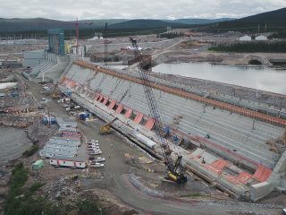 A large hydroelectric dam under construction, with cranes, vehicles, and equipment along the site; water on one side and construction trailers and buildings nearby under a cloudy sky.