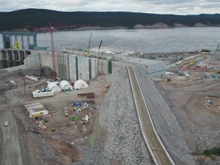 Aerial view of a large hydroelectric dam construction site beside a river, with cranes, construction equipment, buildings, and rocky embankments surrounded by forested hills.