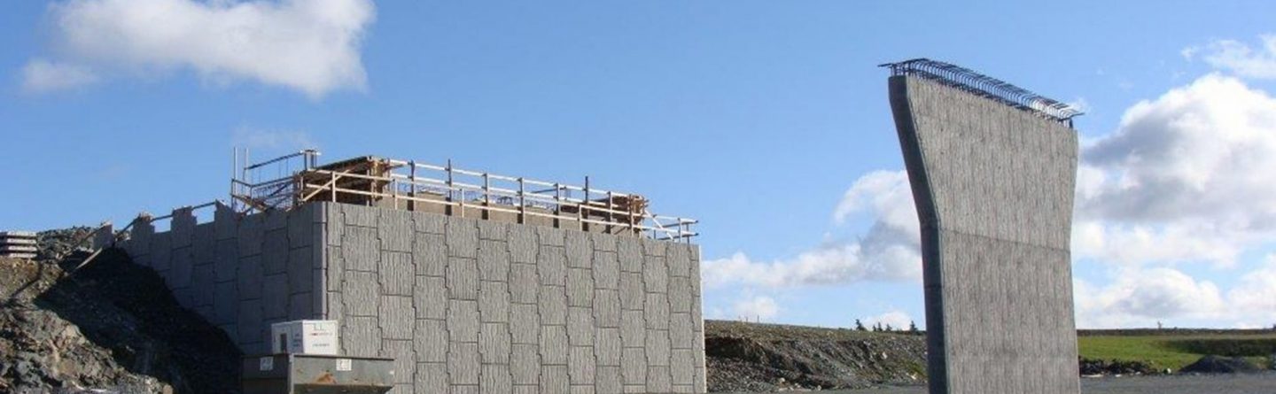 Two concrete bridge abutments stand on a gravel lot; one is tall and rectangular with wooden scaffolding, and the other is curved, standing separately under a blue sky with scattered clouds.