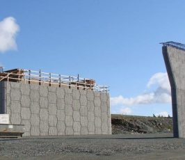 Two concrete bridge abutments stand on a gravel lot; one is tall and rectangular with wooden scaffolding, and the other is curved, standing separately under a blue sky with scattered clouds.