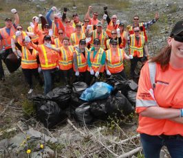 A group of people wearing orange safety vests and hats pose outdoors with garbage bags full of collected litter, celebrating a successful clean-up event; a smiling woman stands in front.