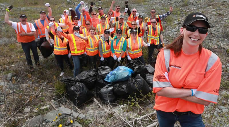 A group of people wearing orange safety vests and hats pose outdoors with garbage bags full of collected litter, celebrating a successful clean-up event; a smiling woman stands in front.