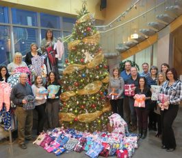 A group of people stands around a decorated Christmas tree on an indoor staircase, holding pajamas. Piles of more pajamas are arranged at the tree’s base. The setting appears festive and cheerful.