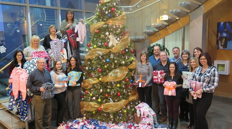 A group of people stands around a decorated Christmas tree on an indoor staircase, holding pajamas. Piles of more pajamas are arranged at the tree’s base. The setting appears festive and cheerful.