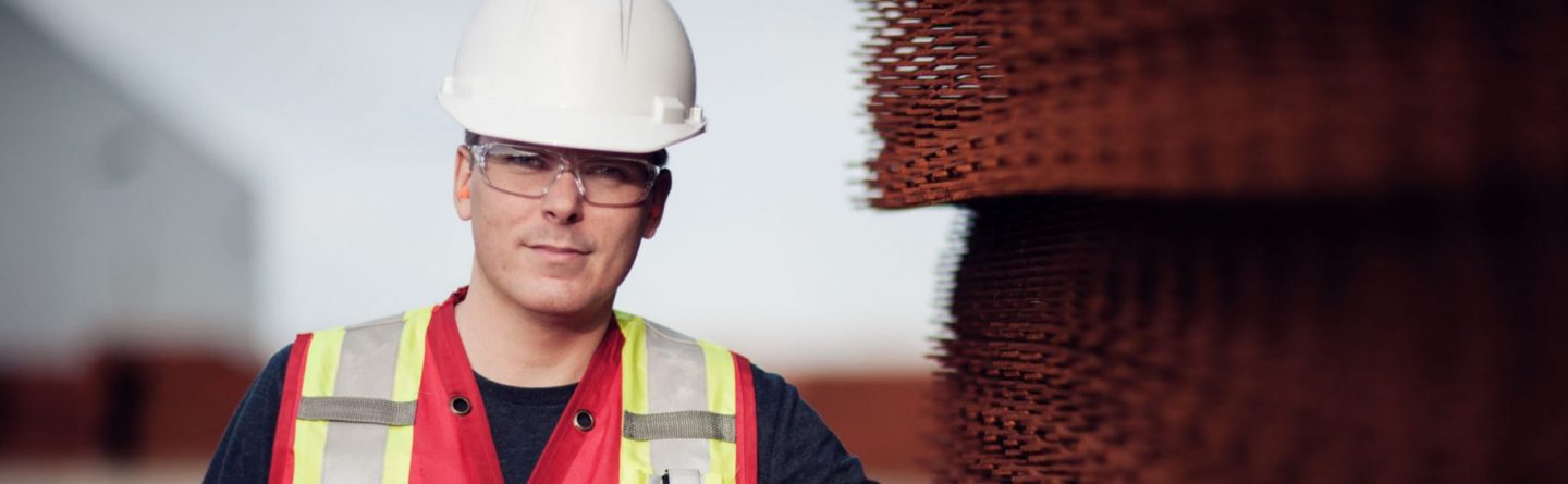 A construction worker wearing a white hard hat, safety glasses, and a reflective vest stands next to stacked metal mesh panels at an outdoor worksite.