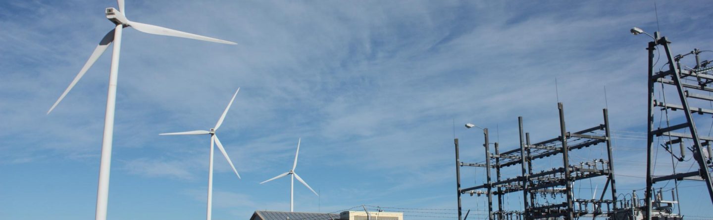 Three large wind turbines stand in the background near a fenced electrical substation and a parked white utility vehicle under a blue sky with wispy clouds.