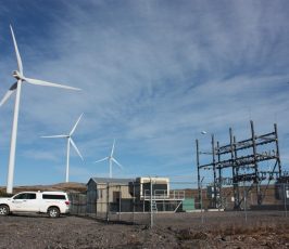 Three large wind turbines stand in the background near a fenced electrical substation and a parked white utility vehicle under a blue sky with wispy clouds.
