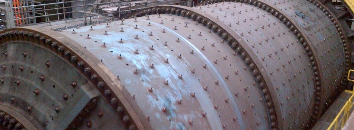 Large industrial ball mill with a cylindrical, metal exterior covered in bolts, situated in a factory setting with structural beams, cables, and railings visible in the background.