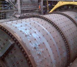 Large industrial ball mill with a cylindrical, metal exterior covered in bolts, situated in a factory setting with structural beams, cables, and railings visible in the background.