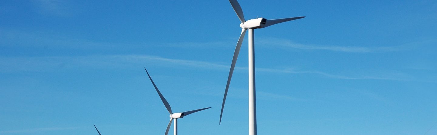 Four large wind turbines stand on rocky terrain under a clear blue sky, generating renewable energy. The landscape is mostly barren except for some low vegetation and rocks.