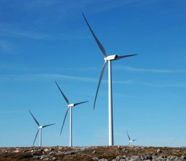 Four large wind turbines stand on rocky terrain under a clear blue sky, generating renewable energy. The landscape is mostly barren except for some low vegetation and rocks.