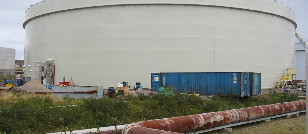 A large, white, cylindrical storage tank is situated outdoors with a blue shipping container and construction materials nearby. Rusted pipes run across the overgrown grassy area in the foreground under a cloudy sky.