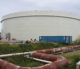 A large, white, cylindrical storage tank is situated outdoors with a blue shipping container and construction materials nearby. Rusted pipes run across the overgrown grassy area in the foreground under a cloudy sky.