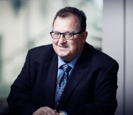 A middle-aged man wearing glasses, a dark suit, blue shirt, and patterned tie sits at a desk, smiling at the camera in an office setting with a blurred background.