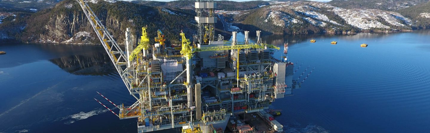 A large offshore oil rig stands in calm blue water near a snow-dusted, forested coastline, with several boats docked alongside the structure under a clear sky.