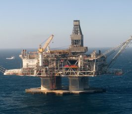 An offshore oil rig stands in the middle of the ocean under a clear sky, with cranes and equipment visible. A supply vessel floats in the distance to the left of the platform.