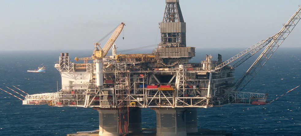 An offshore oil rig stands in the middle of the ocean under a clear sky, with cranes and equipment visible. A supply vessel floats in the distance to the left of the platform.