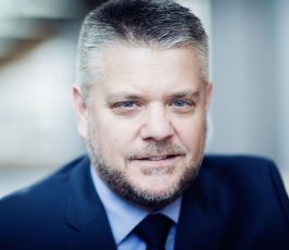 A middle-aged man with short gray hair and a beard, wearing a dark blue suit, light blue shirt, and dark tie, smiles slightly while looking at the camera in a softly blurred indoor setting.