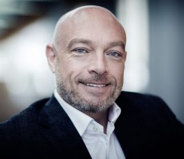 A bald man with a trimmed beard, wearing a dark suit jacket and white shirt, smiles warmly at the camera in a softly lit, modern indoor setting.