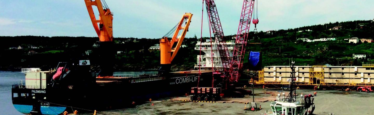 A large cargo ship is docked at a port with two orange cranes unloading materials. A blue and white tugboat is moored nearby. Shipping containers and industrial equipment are on the dock, with hills in the background.