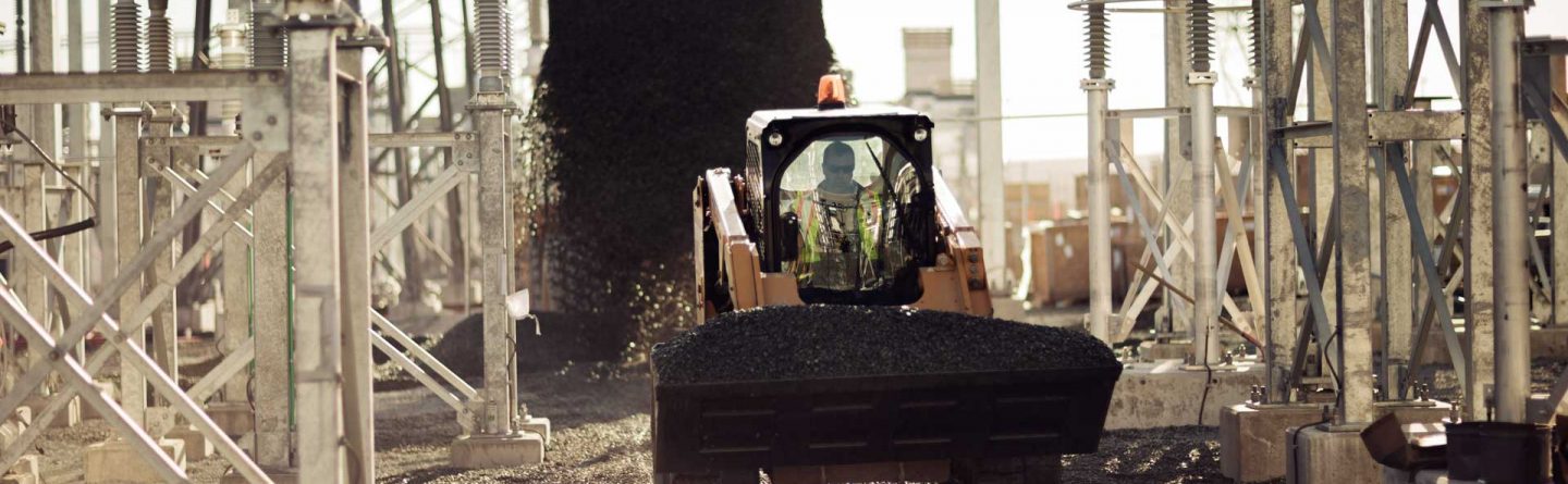 A construction worker operates a small bulldozer, spreading gravel between metal structures at an industrial site during daylight.