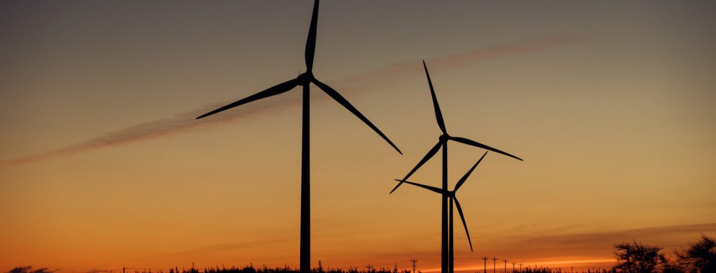 Two wind turbines stand on a hilltop at sunset, silhouetted against an orange and purple sky. A car with headlights on is visible on a nearby road in the lower left corner.